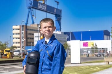 A boy in a blue Babcock boiler suit holding a black welding mask outside, with the Babcock crane visible behind.
