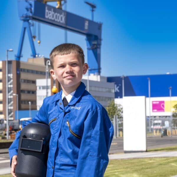 A boy in a blue Babcock boiler suit holding a black welding mask outside, with the Babcock crane visible behind.