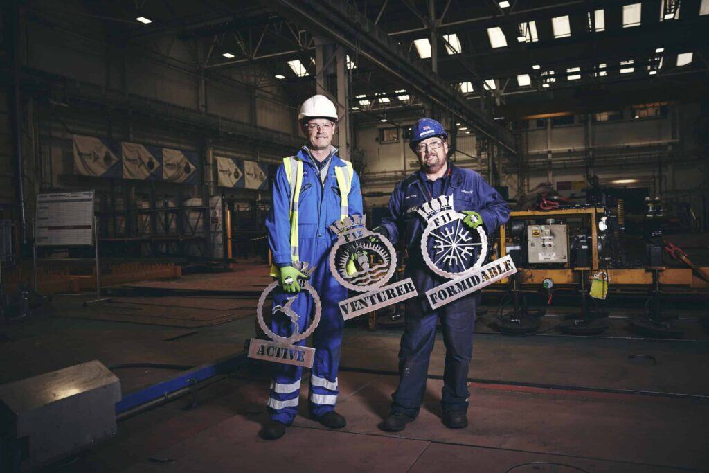 Two Babcock employees in blue overalls holding the crests for each of the three Type 31 frigates - Active